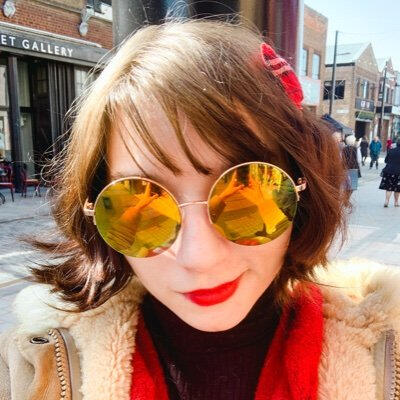 A photograph of Errol Seymour, a person with dark, chin-length hair, red lipstick, and big mirrored sunglasses.